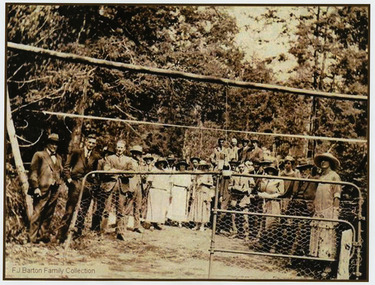 Shows a crowd of people standing behind a gate on a dirt road. In the foreground of the photograph is a log from which is suspended a bottle of champagne. The men and women are all dressed in fine clothes with all women wearing hats.