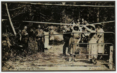 Shows a crowd of people standing behind a gate on a dirt road watching one lady swing a champagne bottle and another cutting a ribbon. The men and women are all dressed in fine clothes with all women wearing hats.