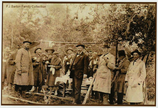 Shows a group of men and women standing around a table eating and drinking. In the background can be seen a log which is hanging between two wooden supports. 