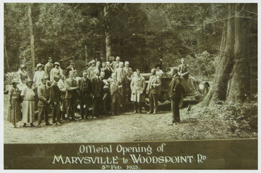 Shows a group of men and women standing and sitting around two cars on a dirt road. The women are all wearing hats.