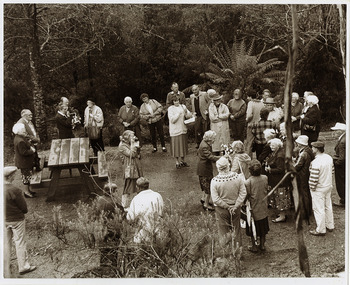 Shows a group of people standing around a wooden picnic bench table talking surrounded by forest.