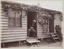 Shows John William Lindt sitting on a chair in the doorway of a weatherboard building looking down at a cat which is lying on the ground in front of the building.