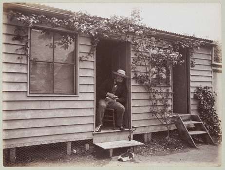 Shows John William Lindt sitting on a chair in the doorway of a weatherboard building looking down at a cat which is lying on the ground in front of the building.