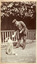 Shows John William Lindt interacting with a dog. In the background is a fence enclosing a vine covered building.