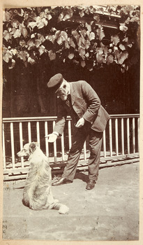 Shows John William Lindt interacting with a dog. In the background is a fence enclosing a vine covered building.
