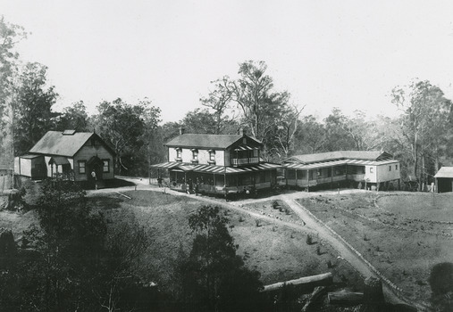 Shows "The Hermitage" at Narbethong in Victoria. Shows a group of three weatherboard buildings, one small and two large. There are several people standing around the buildings. In the foreground are the beginnings of gardens and the buildings back onto forest.