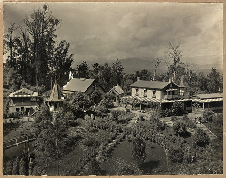 Shows "The Hermitage" at Narbethong in Victoria. Shows a group of weatherboard buildings set amidst extensive gardens. In the left of the photograph there is also a wooden bridge that leads to a small turreted building. At the rear of the main building is another turret. There is a man, woman and a dog walking along a path through the garden. In the background is forest and there are mountains in the distance.