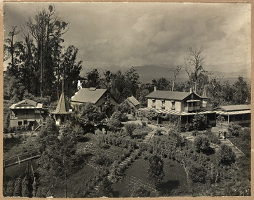 Shows "The Hermitage" at Narbethong in Victoria. Shows a group of weatherboard buildings set amidst extensive gardens. In the left of the photograph there is also a wooden bridge that leads to a small turreted building. At the rear of the main building is another turret. There is a man, woman and a dog walking along a path through the garden. In the background is forest and there are mountains in the distance.