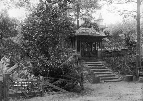 Shows the elaborate entrance gate to "The Hermitage" in Narbethong in Victoria. Shows a flight of steps leading up to a small gatehouse. At the left of the photograph is a wooden and wire fence behind which are tree ferns and trees. At the right of the photograph is a large tree with a wooden ladder leaning against it. In the background can be seen the main building that is "The Hermitage".
