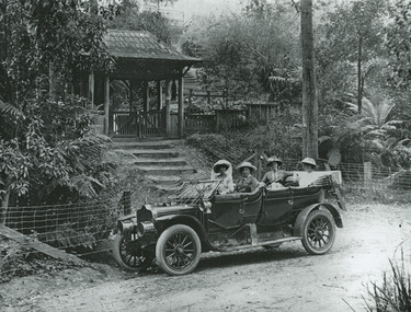 Shows a group of four women in an open tourer car outside the entrance gatehouse to "The Hermitage" at Narbethong in Victoria. Shows a flight of stairs leading up to the gatehouse.