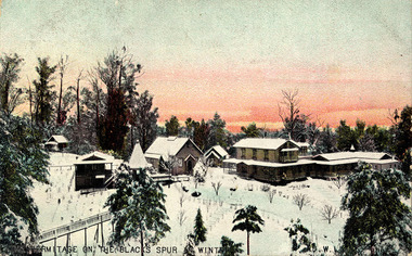 Shows the buildings that are "The Hermitage". Shows a group of four large buildings in addition to one small building. One building has a turret attached and there is a wooden bridge in the left of the photograph that leads to another turret. In the foreground are extensive gardens all covered in snow. The background is forested with mountains in the distance. The title of the postcard is along the lower edge.