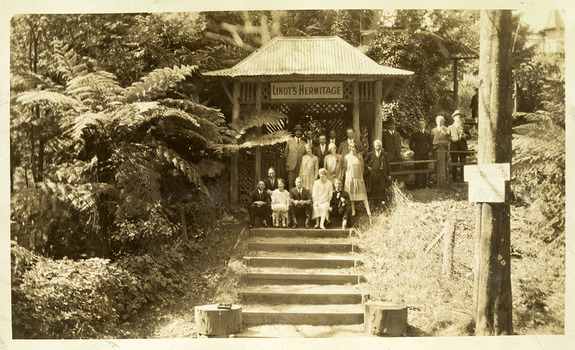 Shows a large group of men and women sitting and standing at the entrance gatehouse at "The Hermitage" at Narbethong in Victoria. There is a sign saying "Lindt's Hermitage handing in the gatehouse.