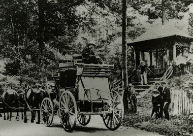 Shows a Cobb & Co coach drawn by four horses standing at the gatehouse at the entrance to "The Hermitage" at Narbethong in Victoria. On the side of the coach can be seen a sign which says MARYSVILLE. The driver is sitting on top of the coach at the rear. There is three men and a boy standing near the coach. There is a man standing with a camera on a tripod on the steps leading up to the gatehouse and there are two other men standing next to the gatehouse.