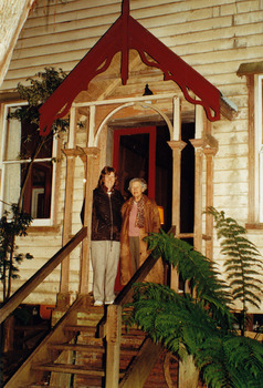 Shows two women standing at the top of some stairs that lead into a weatherboard building. The two women are standing underneath an elaborate timber portico.