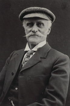 Shows a portrait of a man, from the waist up, seated facing to right with head turned to front. The man has a mustache and is wearing a hat.
