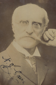 Shows a portrait of a man who is facing the camera. His left hand is holding his head in position and he has a mustache and is wearing glasses. His signature is across the left hand lower corner of the photograph.