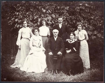 Shows a group of seven people, five women and two men posing in a garden. Nicholas John Caire is seated in the middle of two women who are also seated, and there are three women and a man standing behind them. 