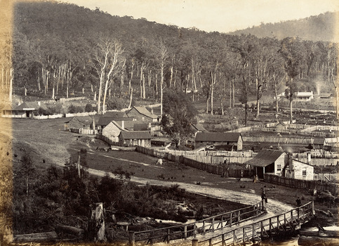 Show the early development of the township of Marysville in Victoria. In the foreground is a wooden bridge. There is a man sitting on the top railing and there is another man standing close by. In the background are a number of fenced off properties, some of which have timber buildings on them. There is also evidence of several trees having been cut down.