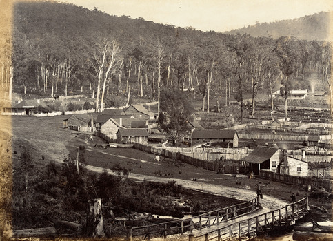 Show the early development of the township of Marysville in Victoria. In the foreground is a wooden bridge. There is a man sitting on the top railing and there is another man standing close by. In the background are a number of fenced off properties, some of which have timber buildings on them. There is also evidence of several trees having been cut down.