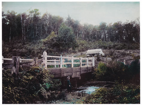 Shows a wooden, white painted bridge that crosses over a river. In the foreground, standing on the bank of the river, is a young girl wearing a hat. In the background is a timber building surrounded by wooden fences. 