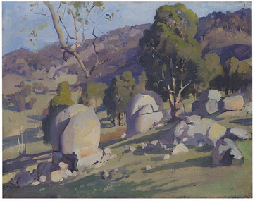 Shows a group of large boulders and smaller rocks on the side of a hill. In the background are several trees and in the distance there are hills and mountains.
