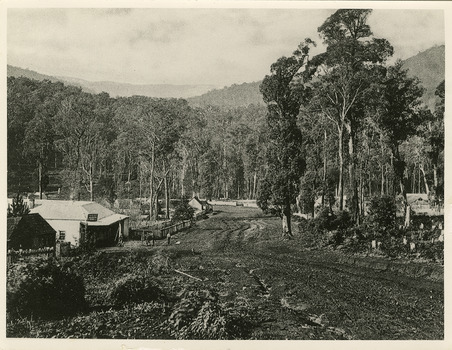 Shows Murchison Street in Marysville in Victoria. Shows a dirt road leading towards a forest. In the left of the photograph is the Keppel's Australian Hotel with four men standing out the front. On the right next to the hotel is another building with a wooden picket fence along its front. On the left of the hotel is fenced off area and further down the street there are more buildings. On the opposite side of the road are more buildings and the road curves around to the left with even more building situated along it.