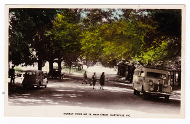 Shows Murchison Street in Marysville in Victoria. Shows a tree lined street with three people crossing the road. In the left of the photograph is two cars, one with a man standing next to it. On the opposite side of the road is a bus with a building in the background. The title of the photograph is along the lower edge of the postcard. On the reverse is a hand written message.