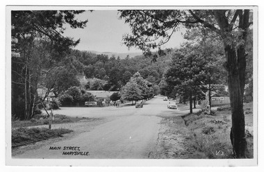 Shows the Main Street in Marysville in Victoria. In the background can be seen the Australia Hotel which became The Cumberland Hotel. Also in the background can be seen a sign with an arrow pointing to the Mary-Lyn Guest House. The title of the photograph is handwritten in black ink on the lower edge. On the reverse of the postcard is a space to write a message and an address and to place a postage stamp. The postcard is unused.