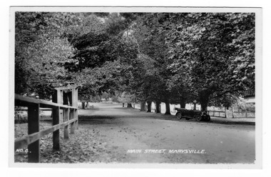 Shows the tree lined main street in Marysville. In the foreground on the left of the photograph can be seen some wooden fencing with an entrance lynch gate. In the right of the photograph can be seen an early model car standing under a tree. The title of the photograph is handwritten in white ink along the lower edge.