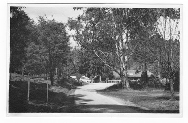 Shows the Marysville-Buxton Road, which leads to Murchison Street in Marysville in Victoria. In the background can be seen The Crossways Hotel with two buses standing outside. In the left of the photograph there is a sign saying 'Marysville' and asking drivers to slow down.