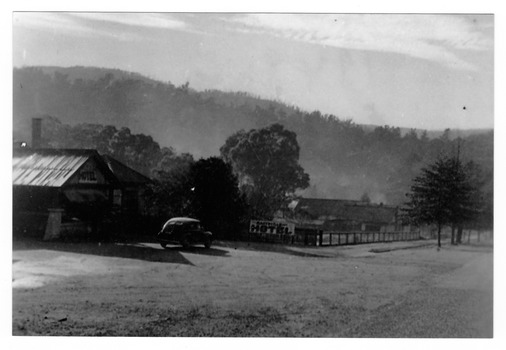 Shows the main street in Marysville in Victoria. In the left of the photograph is The Australian Hotel and further down the road is The Cumberland Guest House. Out the front of The Australian Hotel there is a car parked.