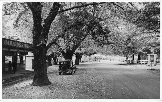 Shows a view of Murchison Street in Marysville in Victoria. Shows the tree lined street with shops on the left hand side of the road. There is a car parked outside the shops. In the right of the photograph is a wooden rail fence with an arbor over an entry way. In the background are three adults and one child walking along the road. 