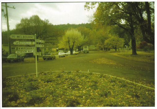 Shows the roundabout at the corner of Murchison and Lyell Streets in Marysville in Victoria. In the foreground there is a signpost with various distances and locations signposted. In the background on the left of the photograph is a building and there are several vehicles parked along the street.