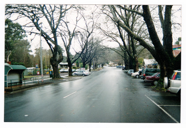 Shows Murchison Street in Marysville in Victoria. Shows the view looking up Murchison Street towards the corner with Pack Road. There are buildings and various vehicles parked on either side of the road and in the foreground is a bus stop shelter. The photograph appears to have been taken after rain.