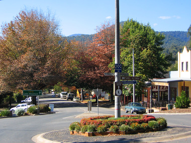 Shows the roundabout at the corner of Murchison and Lyell Streets in Marysville in Victoria. In the right of the photograph there is a row of buildings including a pizza shop. There are cars parked on either side of the street. In the roundabout is a light pole with signs giving directions to various destinations and tourist information. In the left of the photograph can be seen a green sign directing tourists to Lake Mountain.