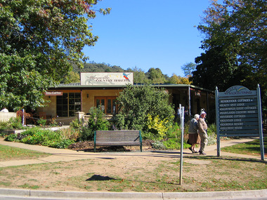 Shows the front of the Marysville Country Bakery in Marysville in Victoria. In the right of the photograph is a wooden sign that informs visitors of the location of various accommodation choices. There is also a wooden bench seat and two people walking on the footpath.