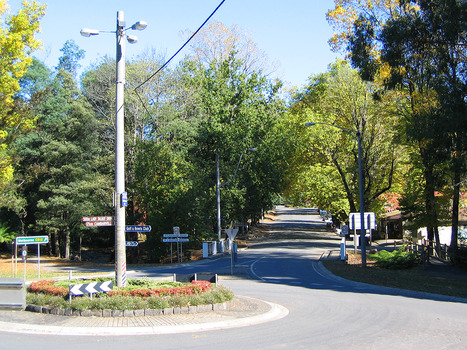 Shows the view looking up Murchison Street in Marysville in Victoria. Shows the view looking up the street from the corner of the Marysville-Buxton Road. In the foreground is a roundabout  with a light pole that has signs to the caravan park, the golf club as well as brown tourist signs to Lady Talbot Drive and Cambarville. There is also a green signing directing people to Melbourne and signs on the F.J. Barton Bridge and Steavenson River.