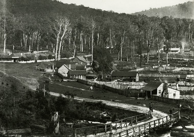 Show the early development of the township of Marysville in Victoria. In the foreground is a wooden bridge. There is a man sitting on the top railing and there is another man standing close by. In the background are a number of fenced off properties, some of which have timber buildings on them. There is also evidence of several trees having been cut down.