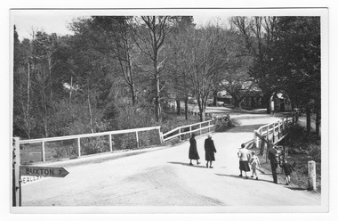 Shows a wooden bridge crossing over a river leading to a road that extends into the town of Marysville in Victoria. Shows two groups of people, including two children, about to walk along the bridge. In the foreground is a sign-post with the distance to Buxton; Healesville is the other destination on the sign-post. In the background, on the right hand side of the street are a series of buildings with cars parked outside them.