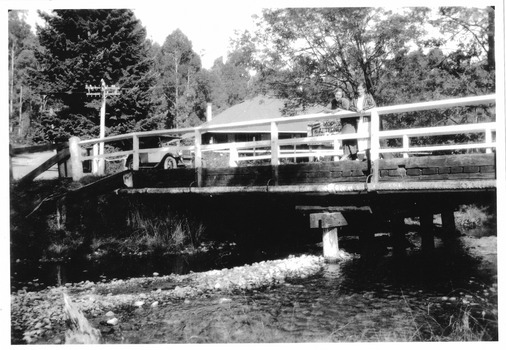 Shows a wooden bridge that crosses over the Steavenson River in Marysville in Victoria. There is two women standing looking over the railing of the bridge with a car parked at the beginning of the bridge. In the background can be seen The Crossways Hotel.