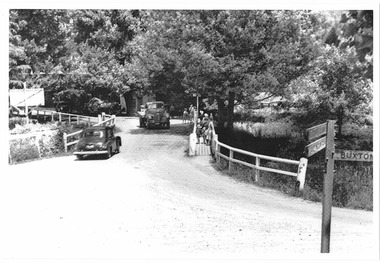 Shows a wooden bridge that crosses over the Steavenson River in Marysville in Victoria. Shows a car and a truck about the cross the bridge. There is a pedistrian walkway along the right hand side of the bridge with people, including children, walking along it. In the foreground is a signpost with various destinations shown.