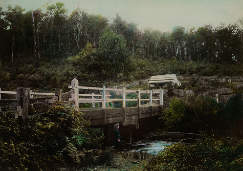 Shows a wooden, white painted bridge that crosses over a river. In the foreground, standing on the bank of the river, is a young girl wearing a hat. In the background is a timber building surrounded by wooden fences. 