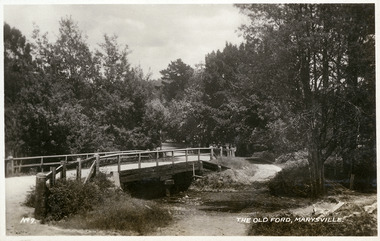 Shows the old ford in Marysville, Victoria. Shows a wooden bridge over the river. In the background the heavily treed road leads off into the distance. The title of the photograph is handwritten in white ink along the lower edge.