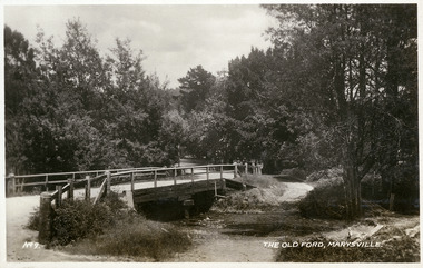 Shows the old ford in Marysville, Victoria. Shows a wooden bridge over the river. In the background the heavily treed road leads off into the distance. The title of the photograph is handwritten in white ink along the lower edge.