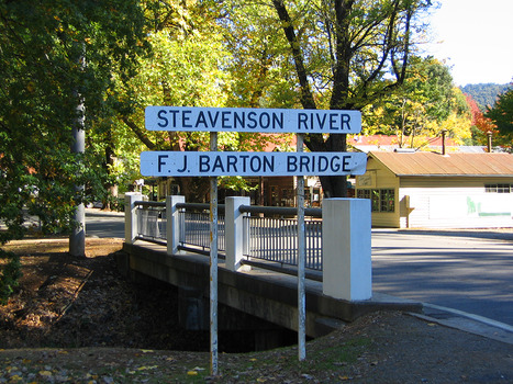 Shows the sign at the F.J. Barton Bridge which crosses the Steavenson River in Marysville in Victoria. In the background is a row of shops.