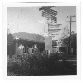 Shows the sign post that stood opposite Pack Road in Marysville in Victoria. Shows the signpost in the foreground with four ladies standing next to it. In the background are two buildings, both shops.