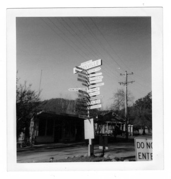 Shows the sign post that stood opposite Pack Road in Marysville in Victoria. In the background are two buildings, both shops.