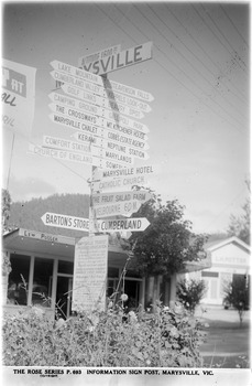 Shows the sign post that stood opposite Pack Road in Marysville in Victoria. In the background are two buildings, both shops.