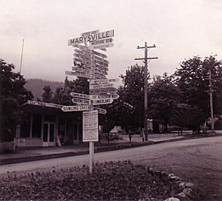 Shows the sign post that stood opposite Pack Road in Marysville in Victoria. Shows various tourist and places of interest sites in and around Marysville and the district. In the background are two buildings, both shops. 