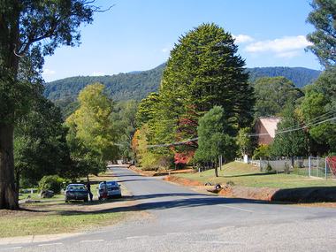 Show the view looking down Sedgwick Street in Marysville in Victoria. In the right of the photograph is a building and in the left are two parked cars with two people standing next to them having a conversation.